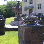 Mignery Sculpture Garden Nebraska Directory Outdoor sculpture garden featuring a central statue of a person on a horse on a stone pedestal with dragon and rider themed statues on brick pedestals and a beige building in the background