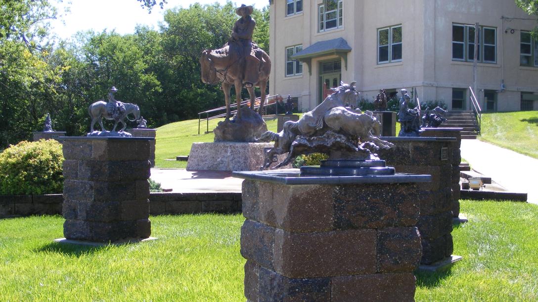 Outdoor sculpture garden featuring a central statue of a person on a horse on a stone pedestal, with dragon- and rider-themed statues on brick pedestals and a beige building in the background.