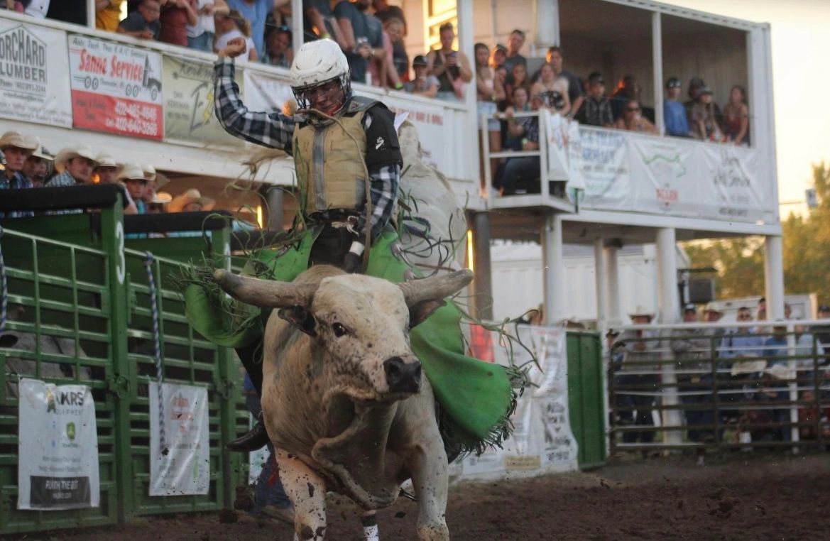 Rodeo bull rider wearing a helmet and vest rides a bucking bull in a muddy arena as spectators watch from the stands.