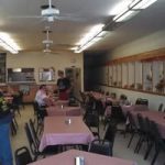 Cafeteria style dining room with pink tablecloths rows of tables and chairs and ceiling fans a few people sit at tables toward the back