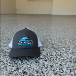 Gray trucker hat with the Modern Garage Coatings logo lying on a speckled gray floor in an empty room