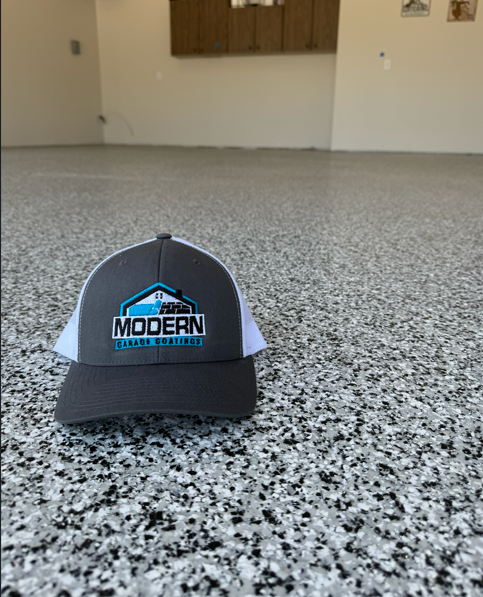 ModernGarageCoatings Gray trucker hat with the Modern Garage Coatings logo lying on a speckled gray floor in an empty room.