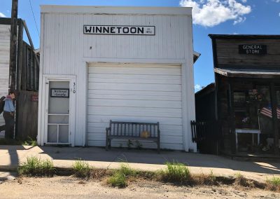 White wooden storefront labeled Winnetoon with a large garage door a wooden bench with a cat and a small side door marked Historical Society on the left a neighboring General Store to the right