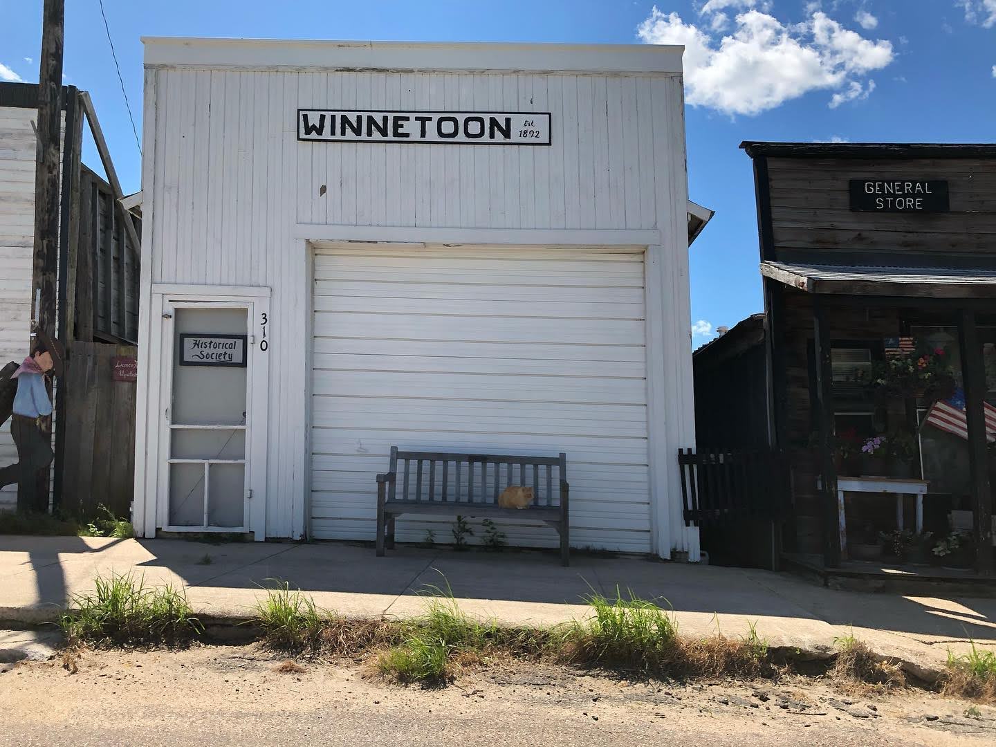 White wooden storefront labeled Winnetoon with a large garage door a wooden bench with a cat and a small side door marked Historical Society on the left a neighboring General Store to the right