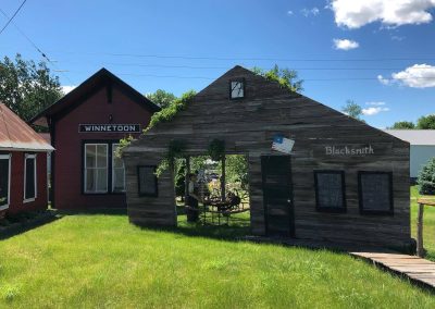 Red wooden building labeled Winnetoon sits beside a weathered blacksmith shop under a bright blue sky green grass in foreground