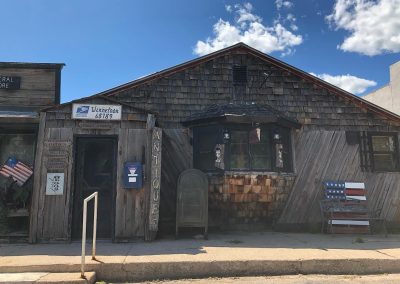 Weathered wooden storefront of an antique shop with eclectic signs and a bay window plus a patriotic flag on the left and a striped bench on the right